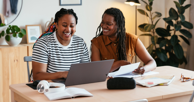 A cheerful teenager and her mother sitting together at a desk, looking at a laptop.